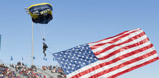 Retired Navy SEAL Jim Woods, aof the U.S. Navy parachute team, the Leap Frogs, presents our nation's flag as he prepares to land during a parachute demonstration as part of Navy Week Fort Worth 2017. Navy Week programs serve as the Navy's principal outreach effort in areas of the country without a significant Navy presence. (Image courtesy of the U.S. Navy by Mass Communication Specialist 3rd Class Danny Kelley)